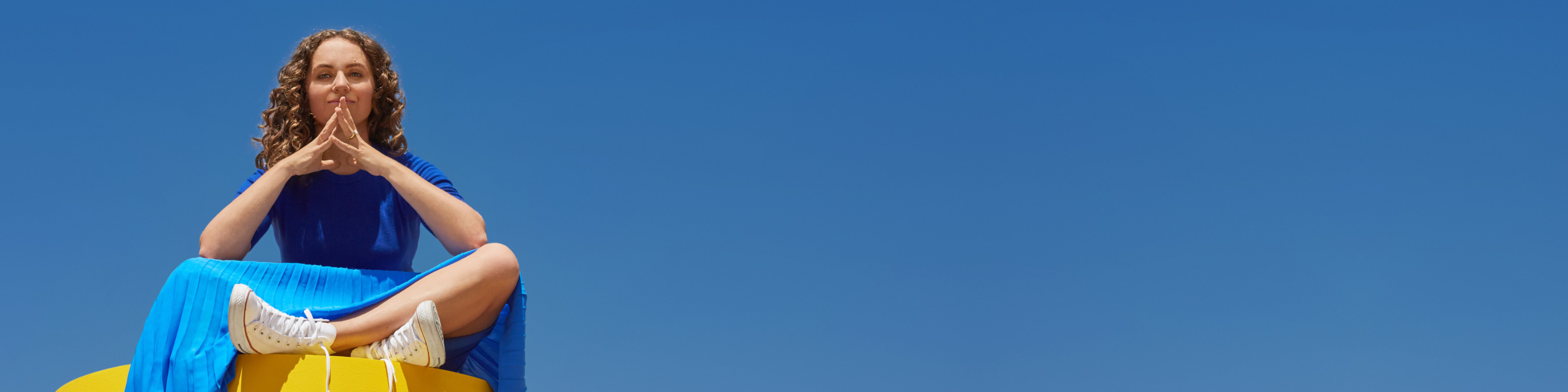 Kat Schneider sitting cross-legged on a yellow object in front of the blue sky.